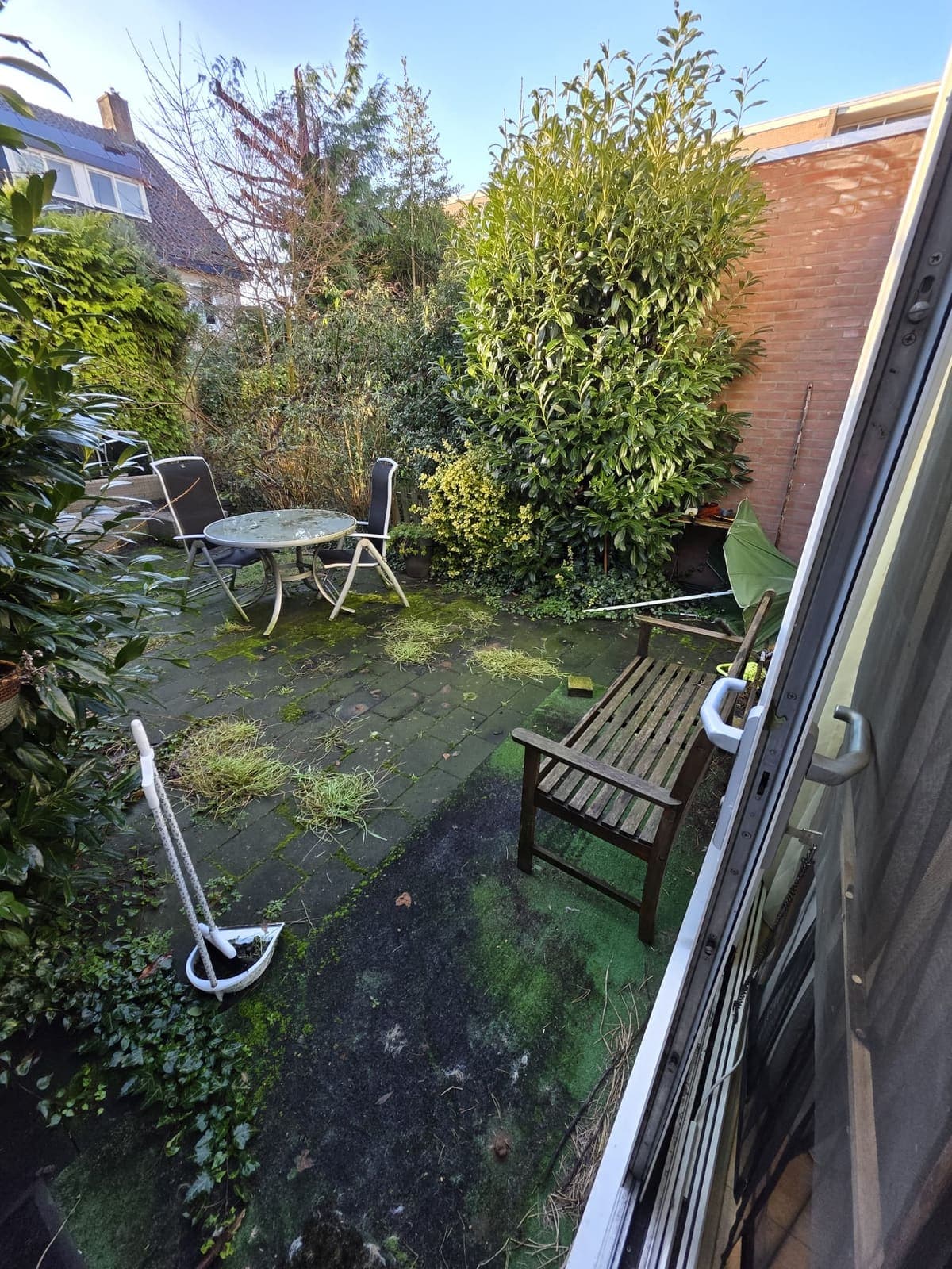 Tuin met mos, onkruid en oude tuinmeubels op de tegels, voordat Buurma Vastgoedservice hem opfriste